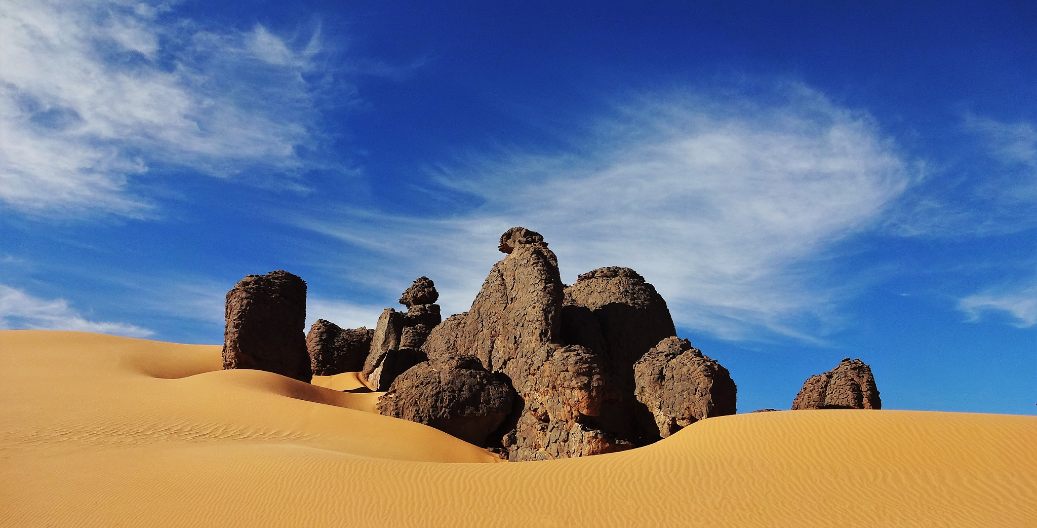 Rochers sculptés par l’érosion au milieu des dunes dorées du Tassili n’Ajjer, sous un ciel bleu.